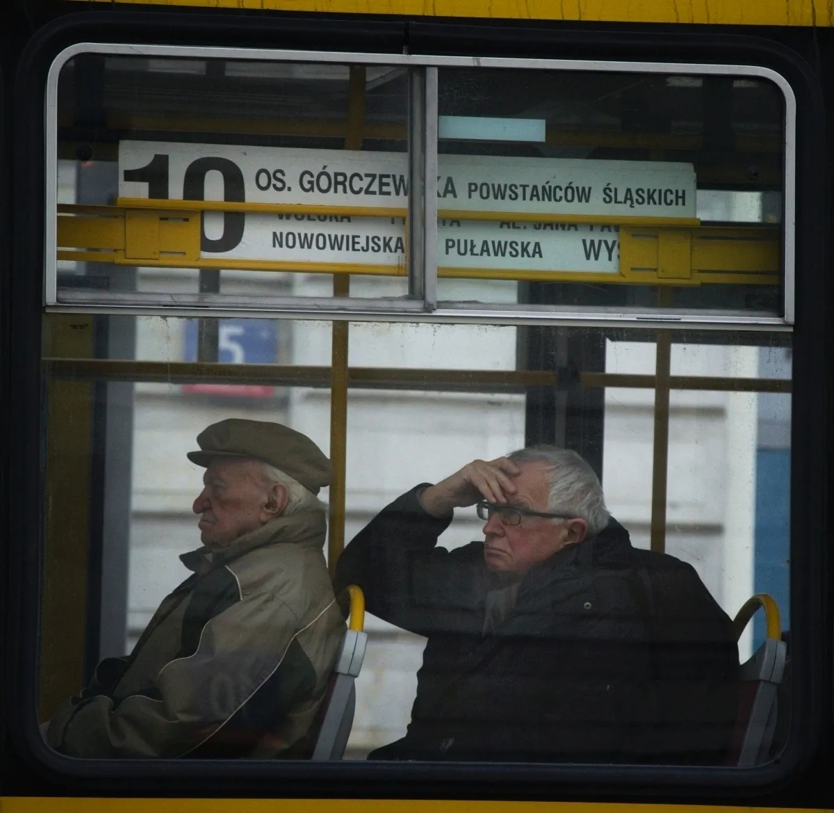 Two older men seated inside a tram, one shading his eyes with his hand